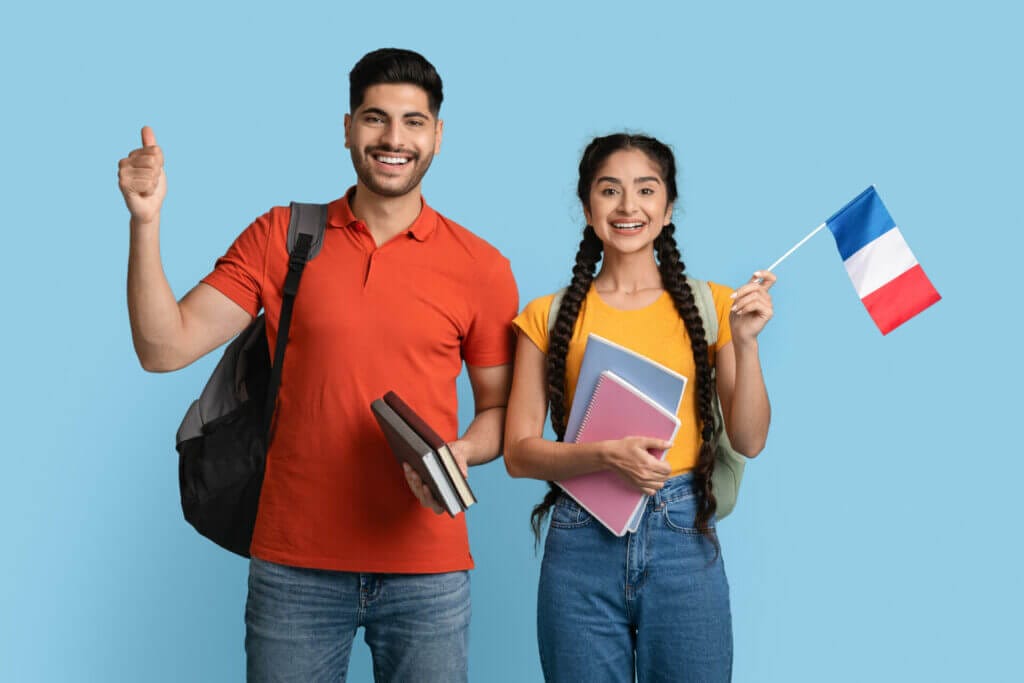 Estudiar en Francia. Hombre y mujer hispanos felices sosteniendo libros de trabajo y la bandera francesa, estudiantes sonrientes con mochilas posando sobre un fondo de estudio azul, recomendando estudiar en el extranjero.